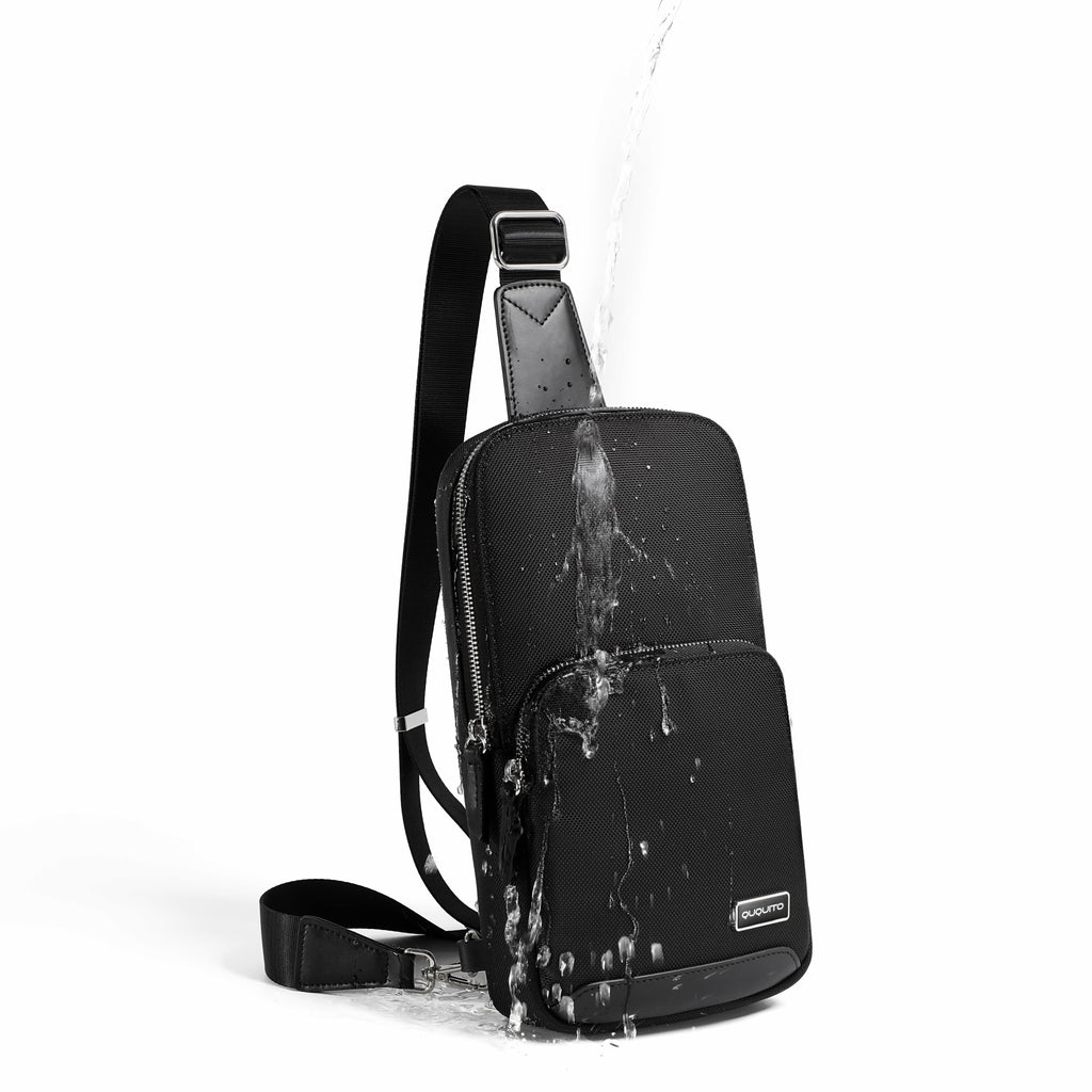 A black sling backpack with a water-resistant finish, shown with water splashing off it against a clean white background.