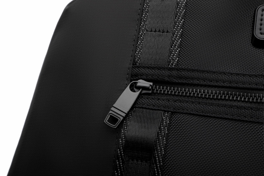 Close-up of a black textured fabric bag with a black zipper and black plastic pull tab, shown against a white background.