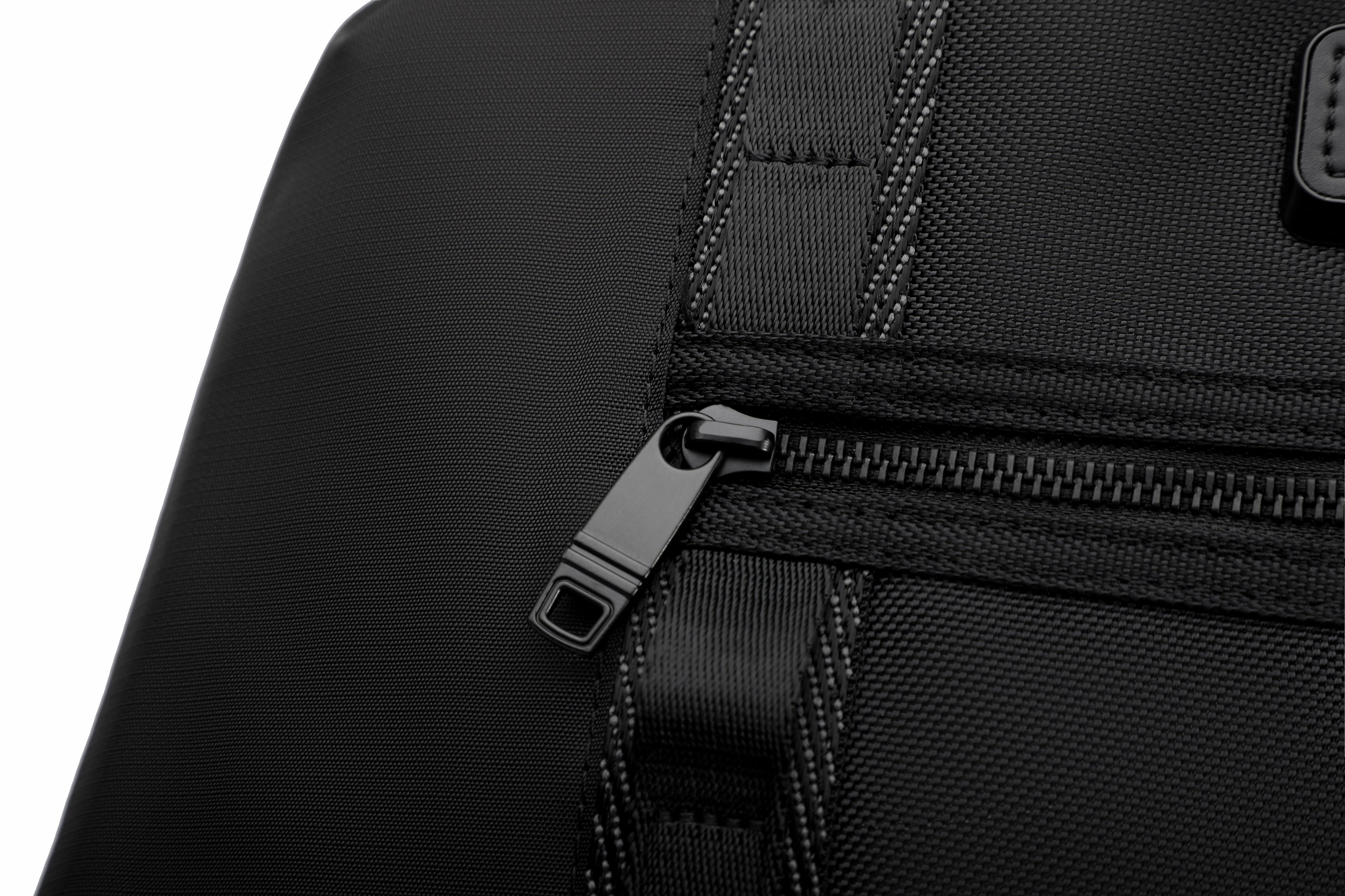 Close-up of a black textured fabric bag with a black zipper and black plastic pull tab, shown against a white background.