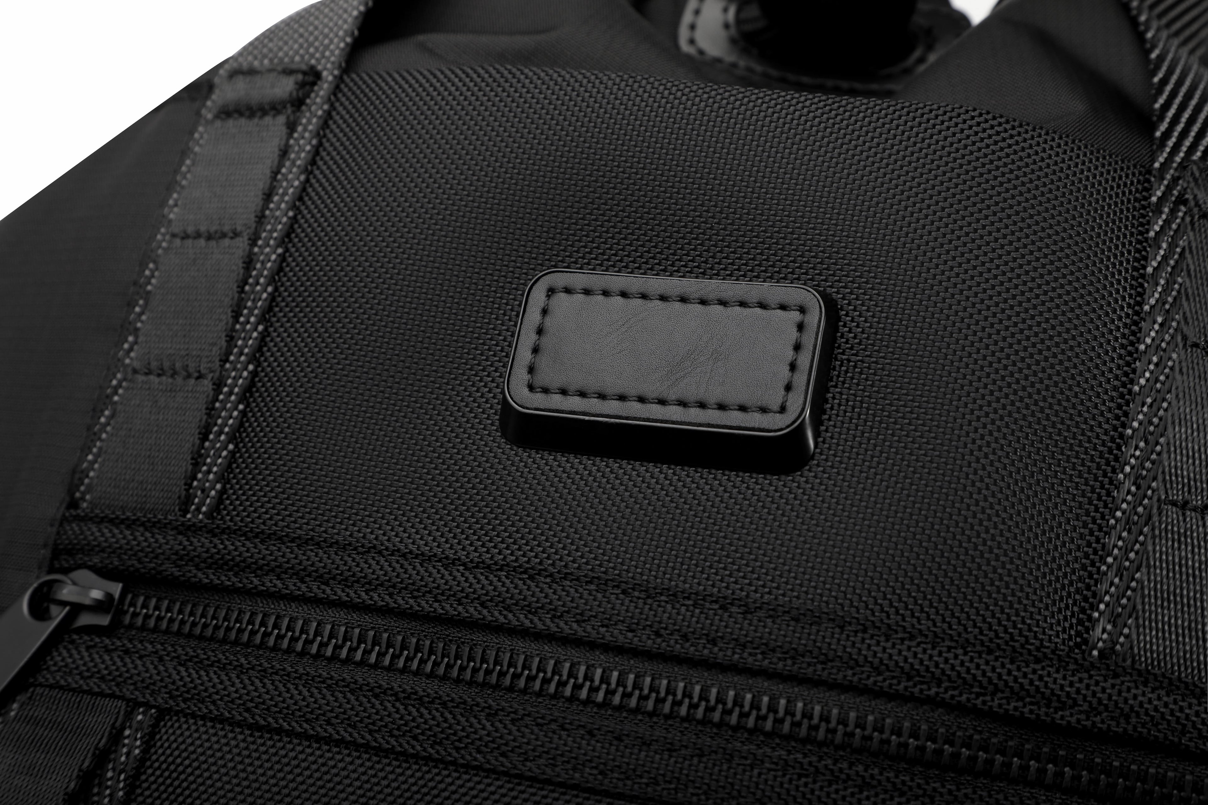 Close-up of a black textured fabric bag with a rectangular black leather patch embossed with the brand name 'DOSHI'.