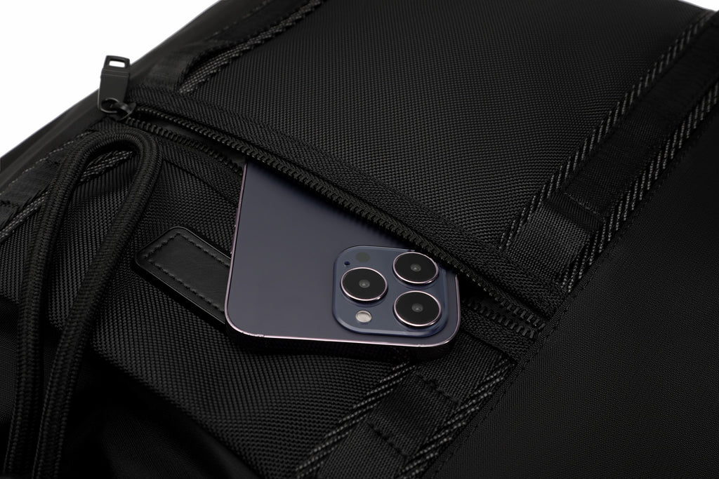 Close-up of a black textured backpack or bag with a zip pocket, showing a dark blue smartphone partially tucked inside.