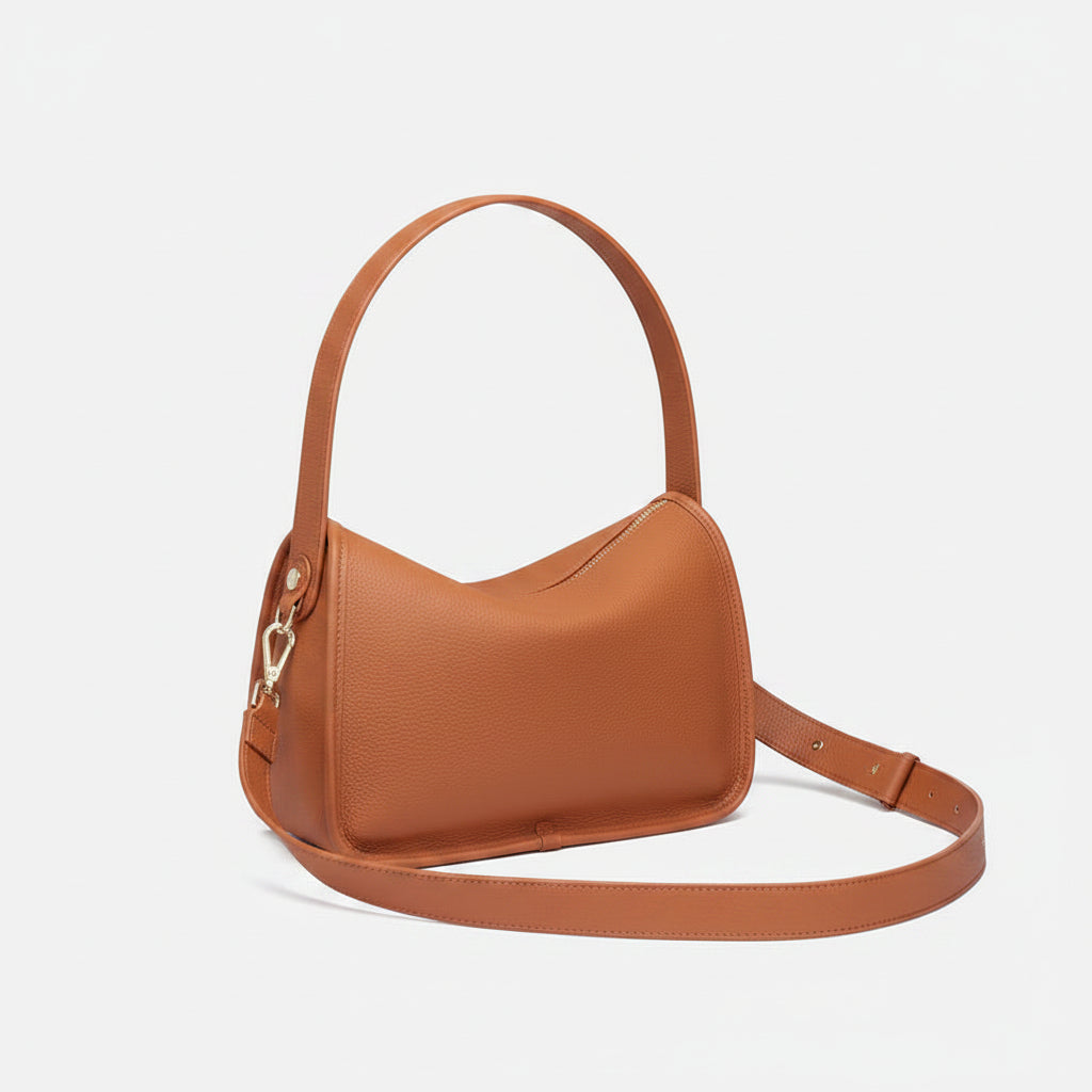 A tan leather shoulder bag with a top handle and a detachable shoulder strap, styled on a white surface with a magazine and a white cup in the background.