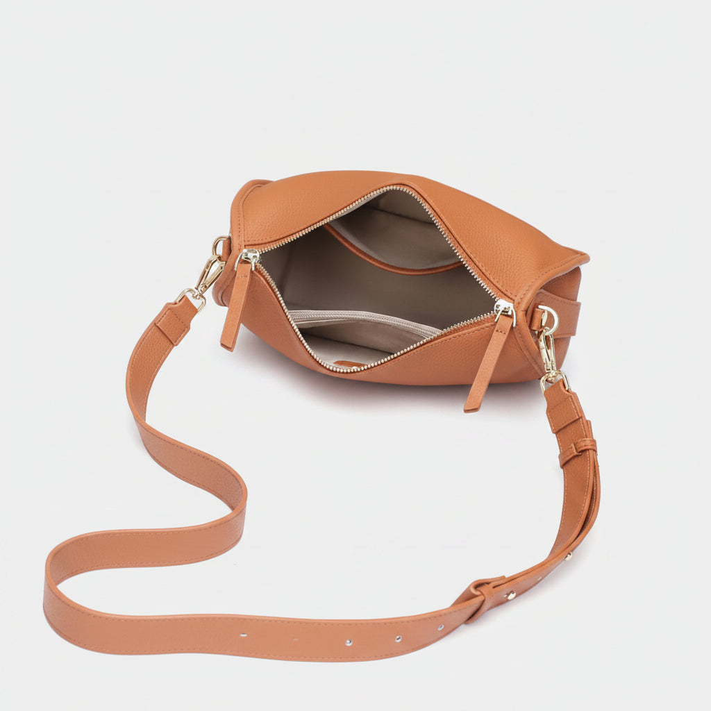 A tan leather shoulder bag with an open zipper, displayed on a white surface with a magazine page underneath. The bag has a matching tan strap and gold-tone hardware.