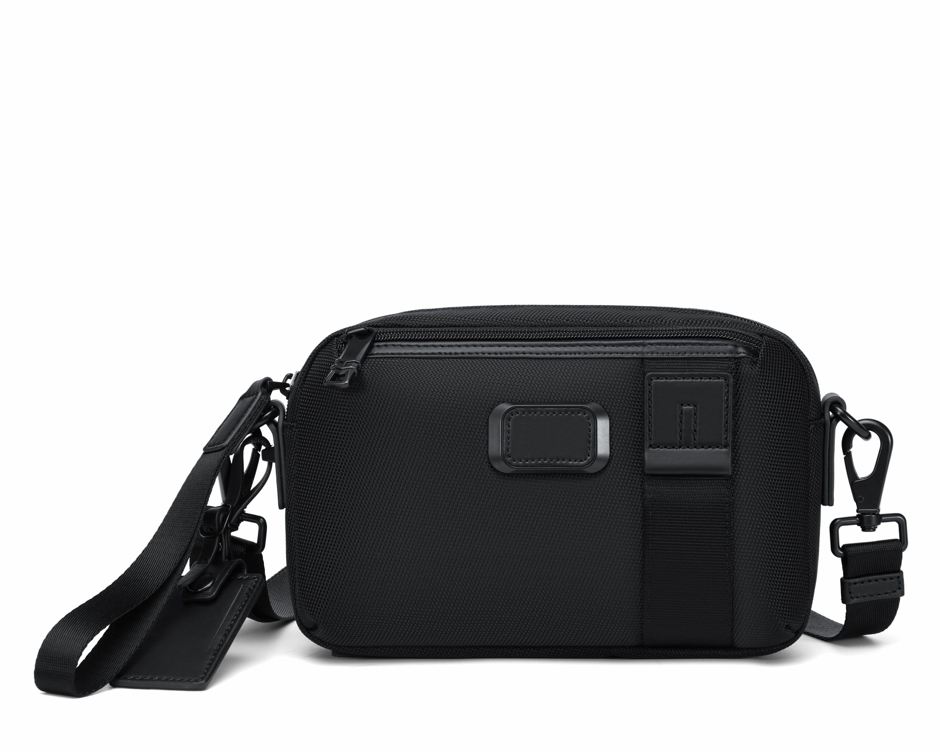 A black rectangular crossbody camera bag with a front zippered pocket, a vertical black strap detail, and a small silver-toned logo plaque, shown on a plain white background.